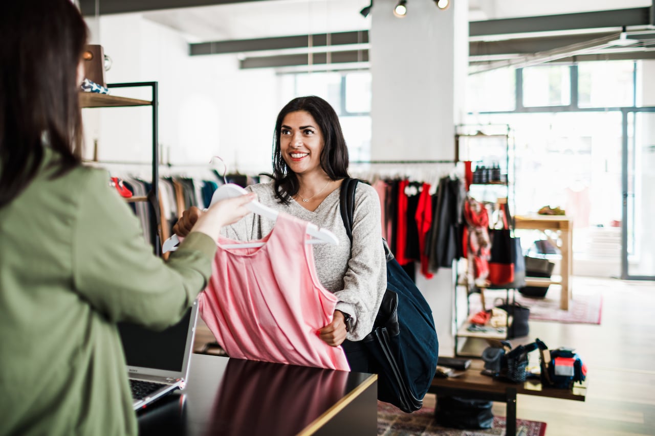 A woman smiling as she is handing a dress to a clothing store cashier so she can pay for it.