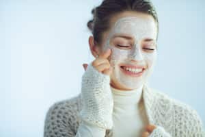 smiling stylish woman in roll neck sweater and cardigan applying white facial mask isolated on winter light blue background.